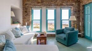 A room with a stone wall, a view of the sea, a white sofa, a teal armchair, and a small wooden table. - Magganari View Villa - Discover Ultimate Summer Happiness! (Magganari Ios Island)