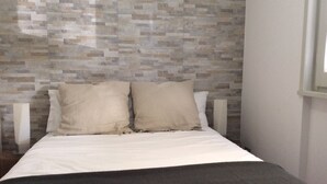 Un lit avec un matelas blanc et deux oreillers beige, placé contre un mur orné d’un motif de mosaïque.