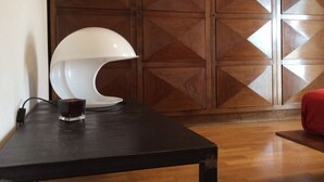Une chambre moderne avec un mur lambrissé en bois, une table noire surmontée d’une lampe blanche, un verre et un coussin rouge.