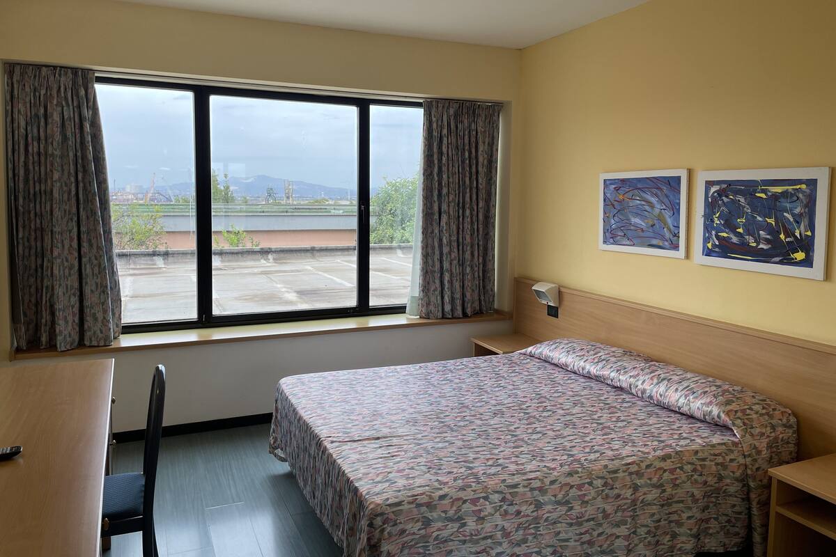 Ein Hotelzimmer mit Bett, Schreibtisch, Stuhl, einem Fenster mit Vorhängen und zwei abstrakten Bildern an der Wand.