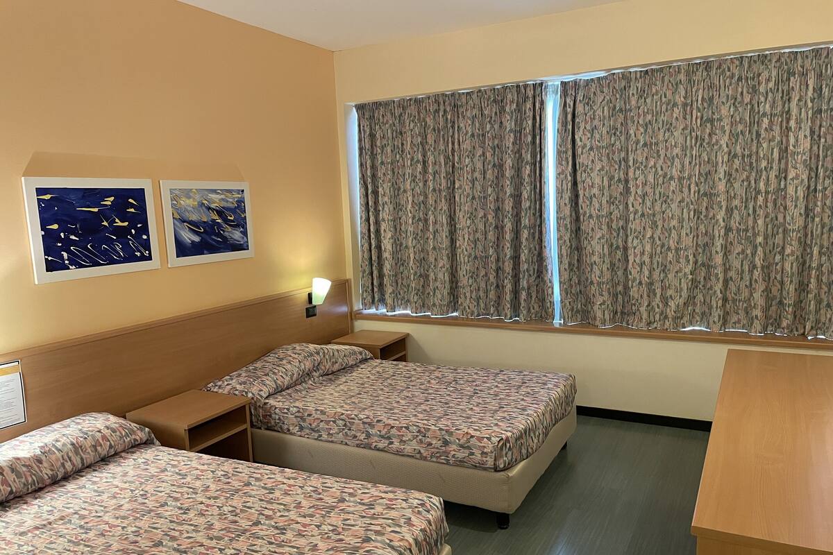 Ein Hotelzimmer mit zwei Betten, einem Nachttisch, einem Schreibtisch und einem Fenster mit Vorhängen.