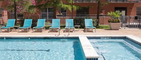 A swimming pool with clear blue water, surrounded by lounge chairs and a brick building with balconies.