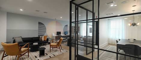 Uma sala de estar moderna com divisória de vidro, um sofá preto, uma cadeira de madeira e uma área de jantar com mesa e cadeiras.