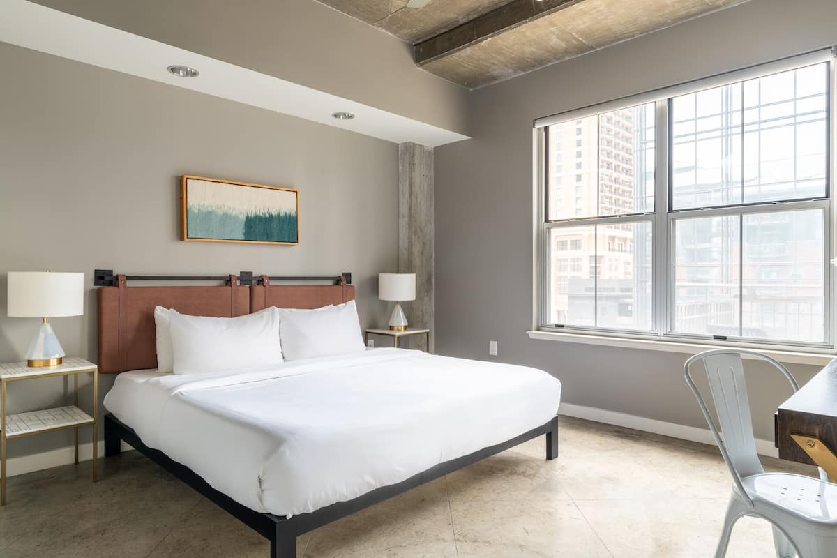 A modern hotel room with a large bed, two bedside tables with lamps, a view of the city through large windows, and a painting on the wall.