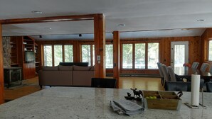 Ett rymligt vardagsrum med en öppen spis i sten, träpanel på väggarna, ett stort fönster och en matplats med bord och stolar.