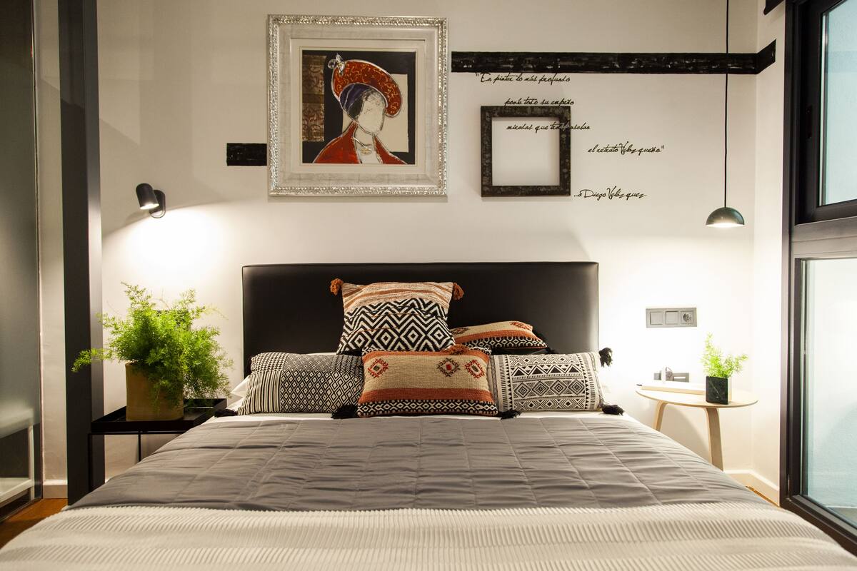 Un dormitorio moderno con una cama grande, almohadillas estampadas, una obra de arte enmarcada y una mesita de noche con una planta.