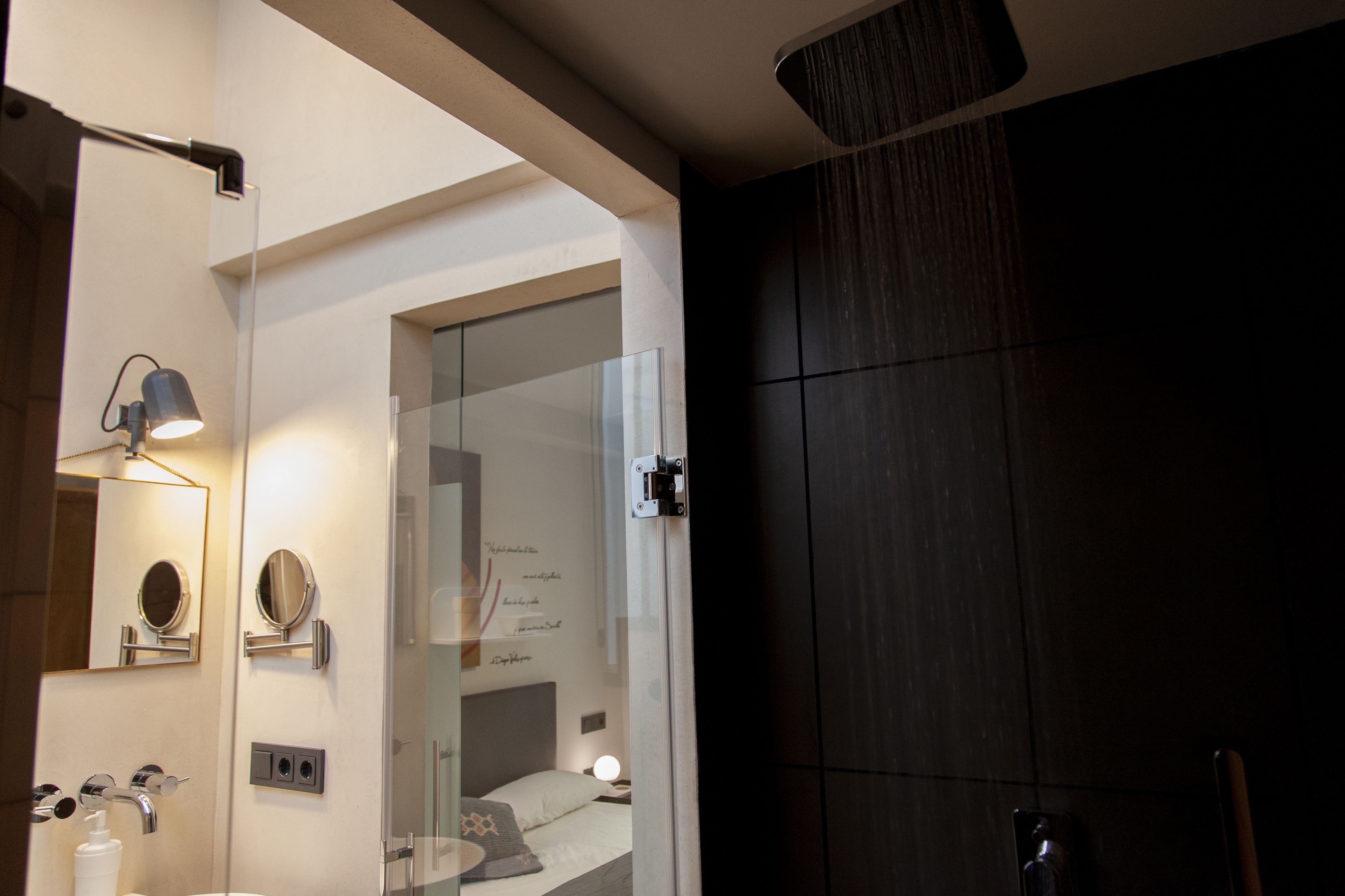 A modern bathroom with a glass shower enclosure, a large mirror, and a bedside table with a lamp.