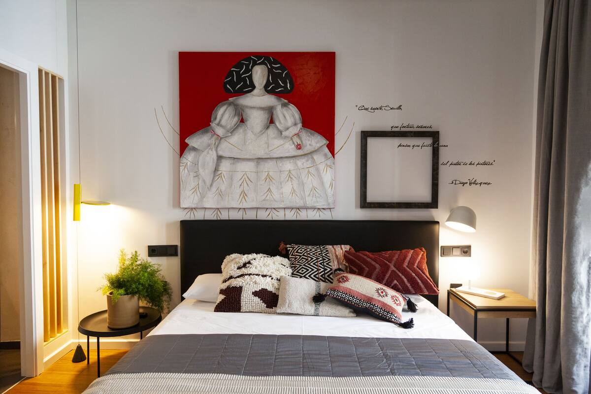 Un dormitorio moderno con una cama grande, varios almohadones, una obra de arte en la pared y una mesita de noche con lámpara.