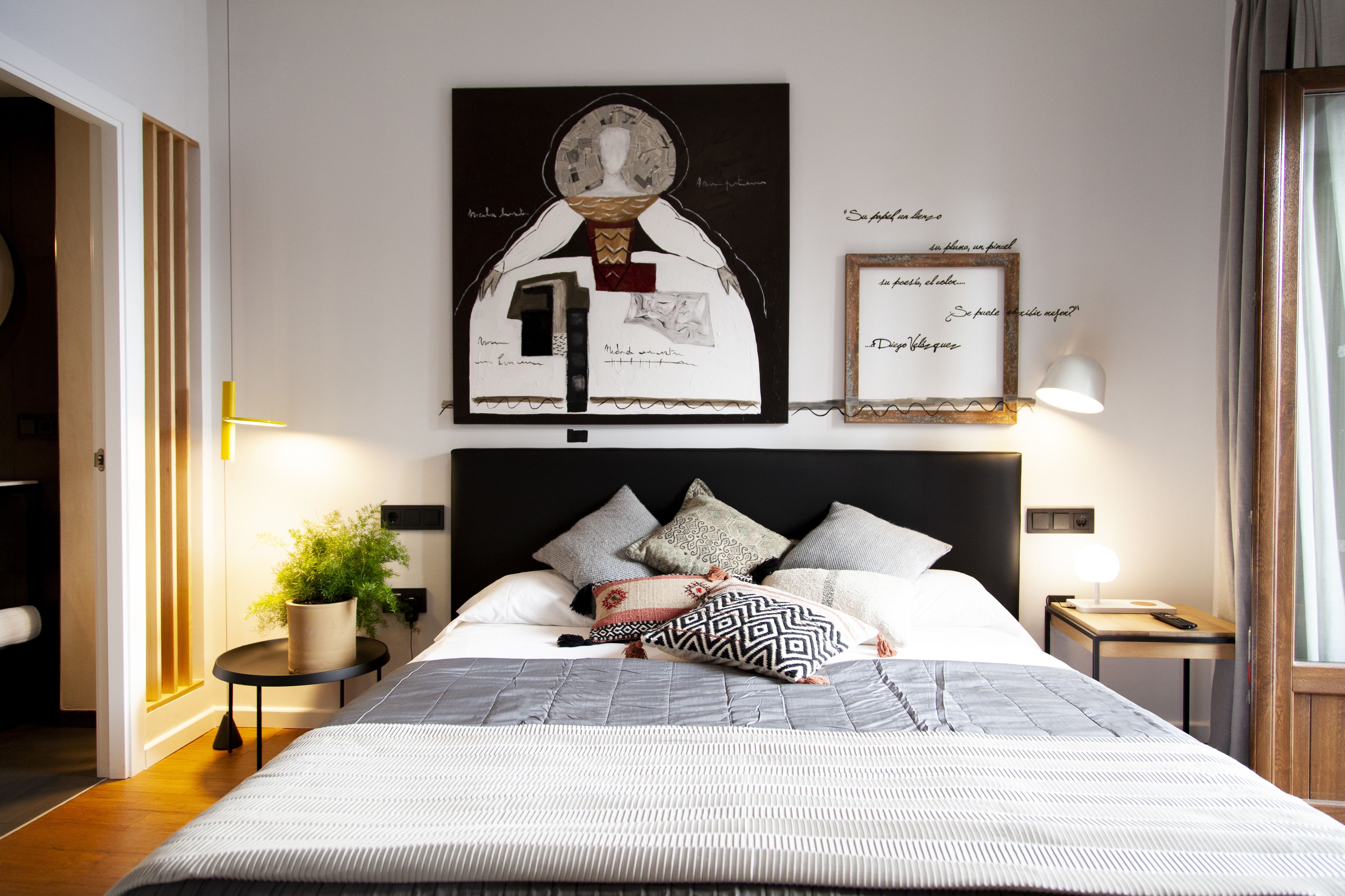 Una cama bien hecha con una colcha estampada, una planta en maceta sobre una mesita de noche y una obra de arte en la pared.