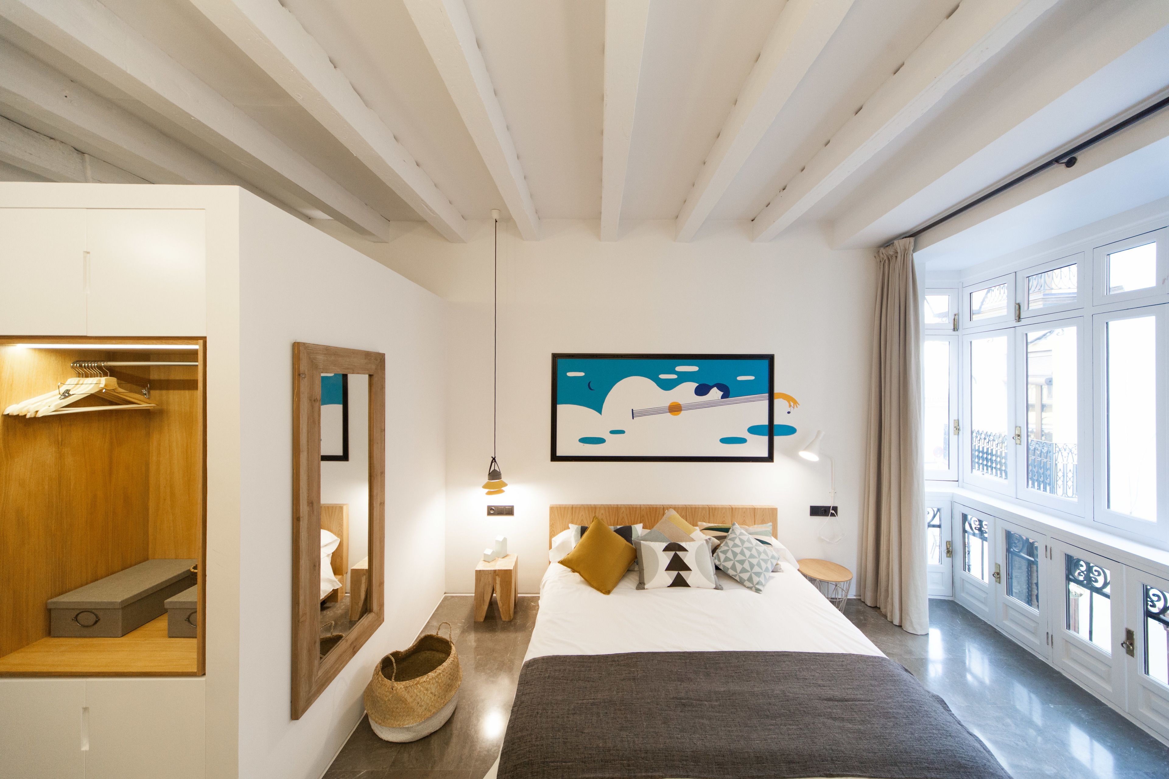 A modern hotel room with a large bed, a wooden wardrobe, a mirror, a painting, and a view of the outside through large windows.