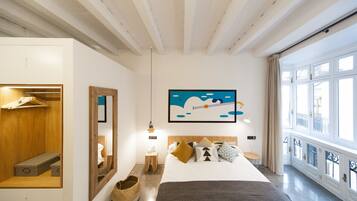 A modern hotel room with a large bed, a wooden wardrobe, a mirror, a painting, and a view of the outside through large windows.