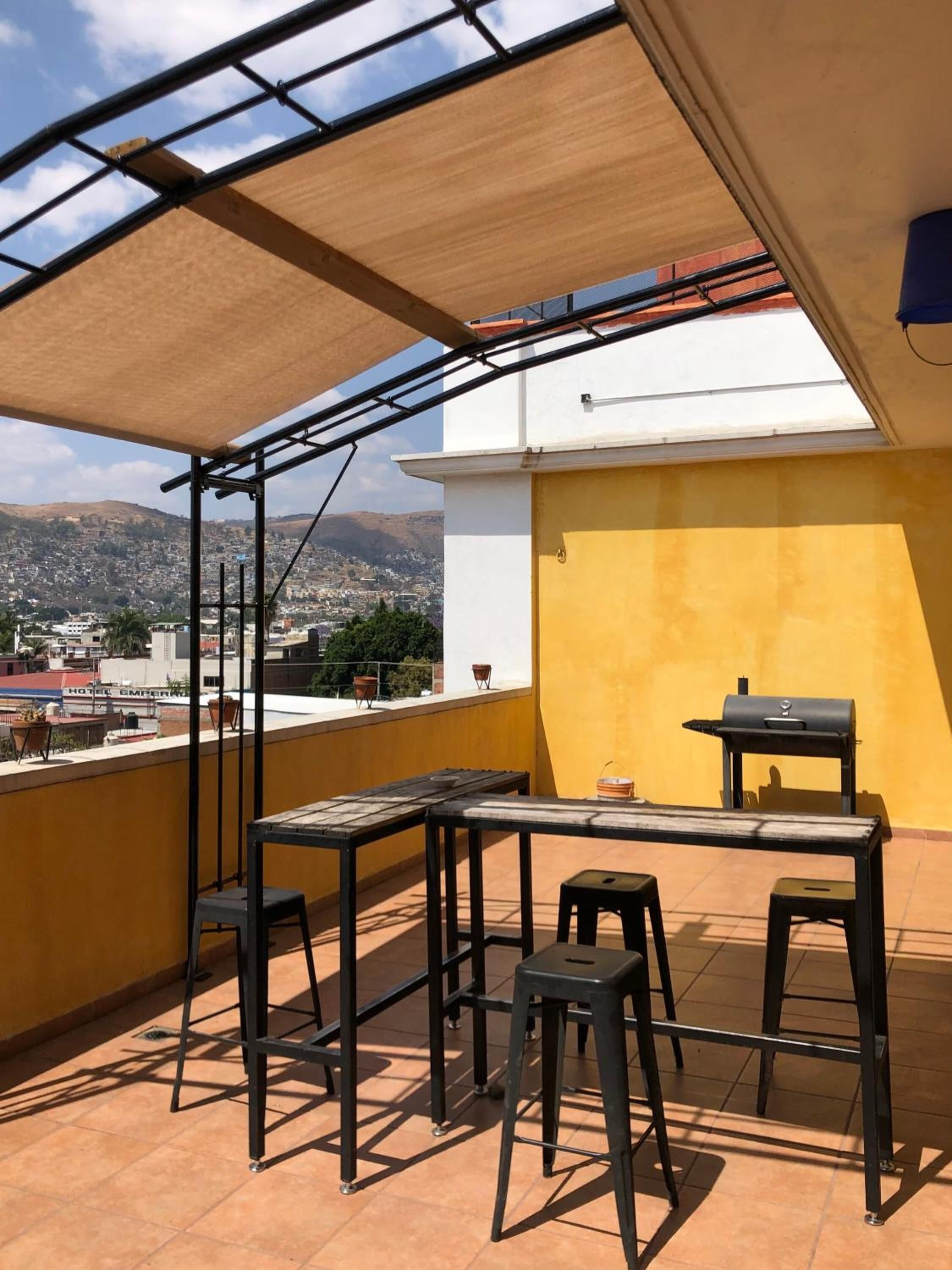 A rooftop terrace with a table and chairs, a grill, and a view of a cityscape and mountains.