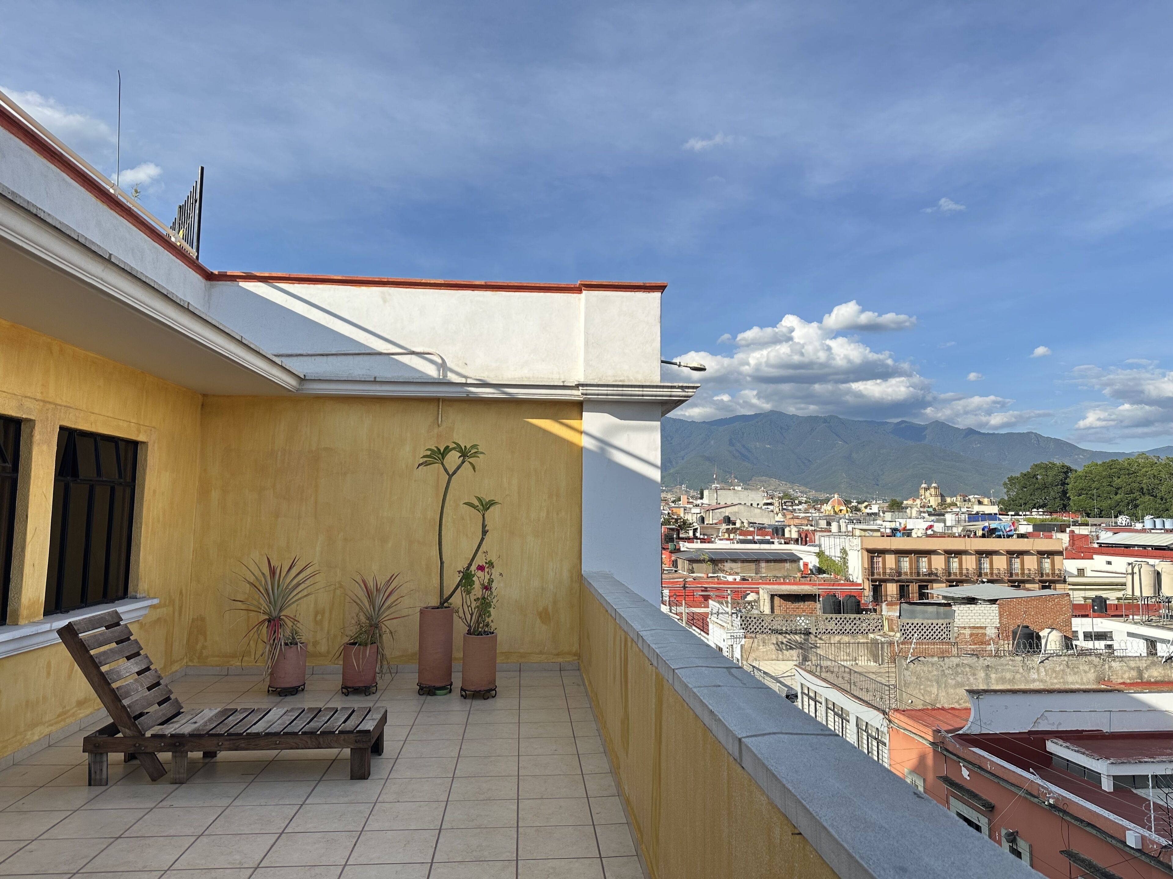 A balcony with a wooden bench, potted plants, and a view of a cityscape with mountains in the background.