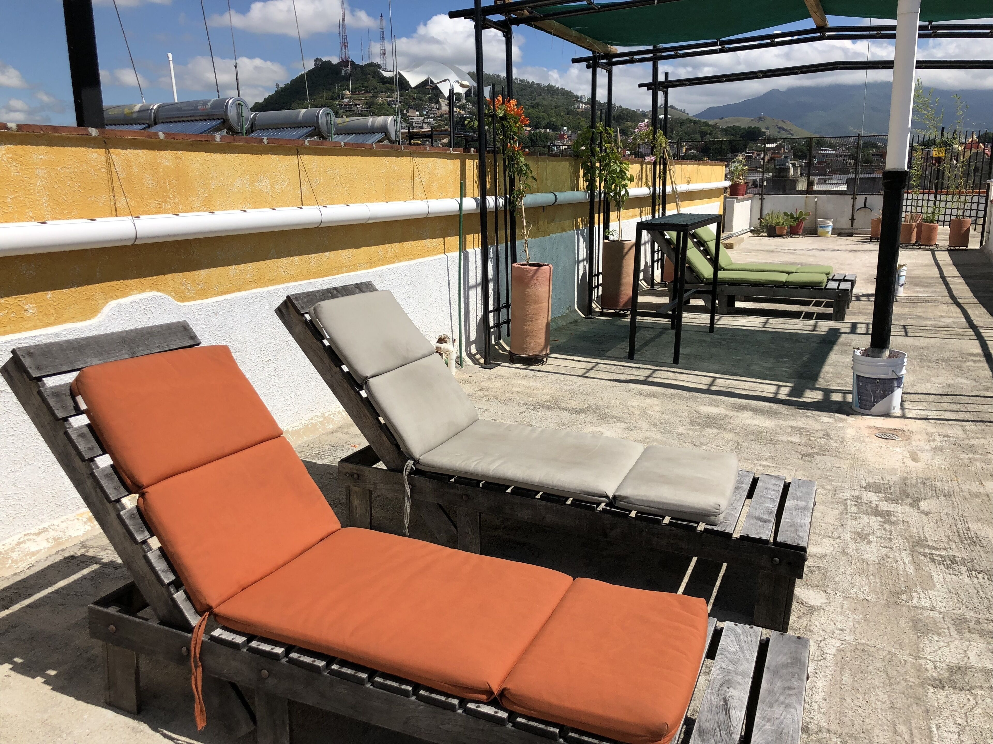 A rooftop terrace with two wooden lounge chairs with orange cushions, a small table, and potted plants.