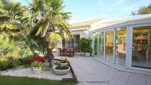 A patio with a dining table and chairs, surrounded by potted plants and a palm tree. - Locatlantique (Aytre)