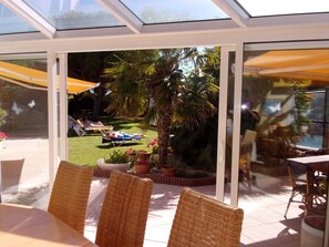 A sunlit patio with wicker chairs and a glass enclosure overlooking a garden with palm trees and a lawn. - Locatlantique (Aytre)