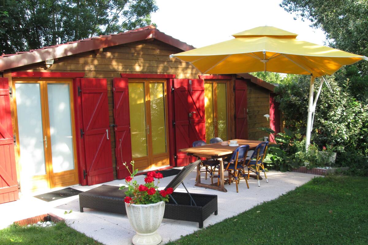 Eine Terrasse mit Holztisch und -stühlen, einem gelben Sonnenschirm und einem Blumentopf mit roten Blüten.