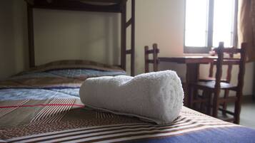Una cama bien tendida con un edredón a rayas, una toalla blanca doblada y una silla de madera en una habitación con ventana y cabecera de madera.