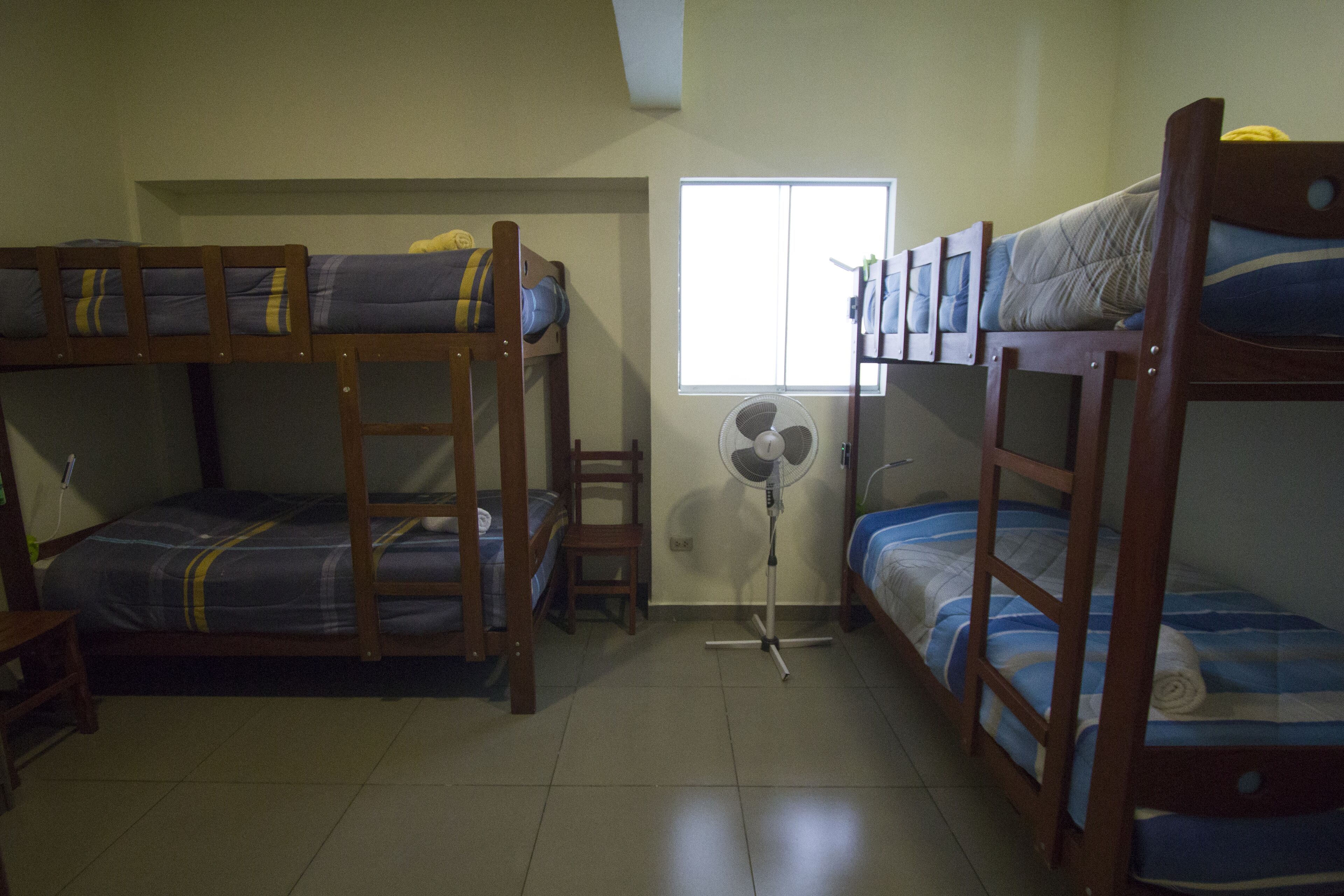A dormitory room with bunk beds, a standing fan, and a window providing natural light.