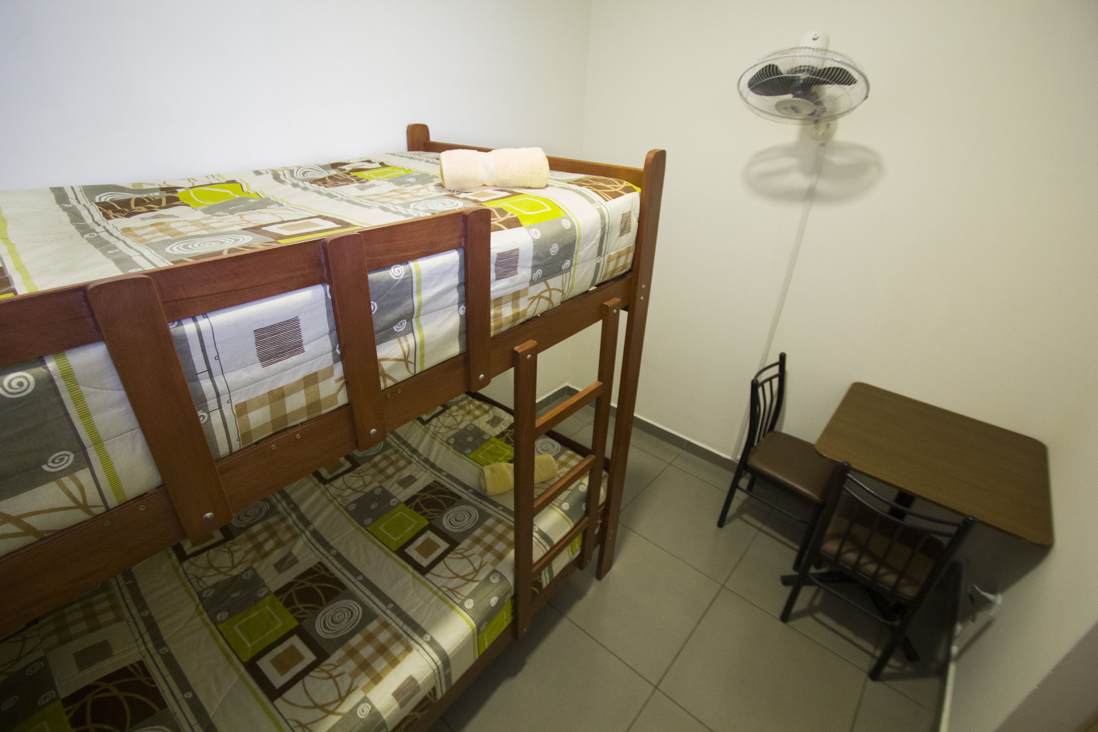 Una cama litera con una colcha estampada, una mesita con una silla y un ventilador de pie.