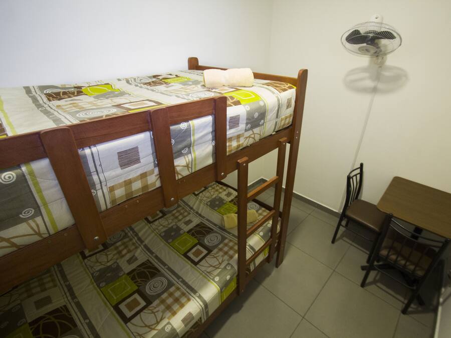Una cama litera con una colcha estampada, una mesita con una silla y un ventilador de pie.