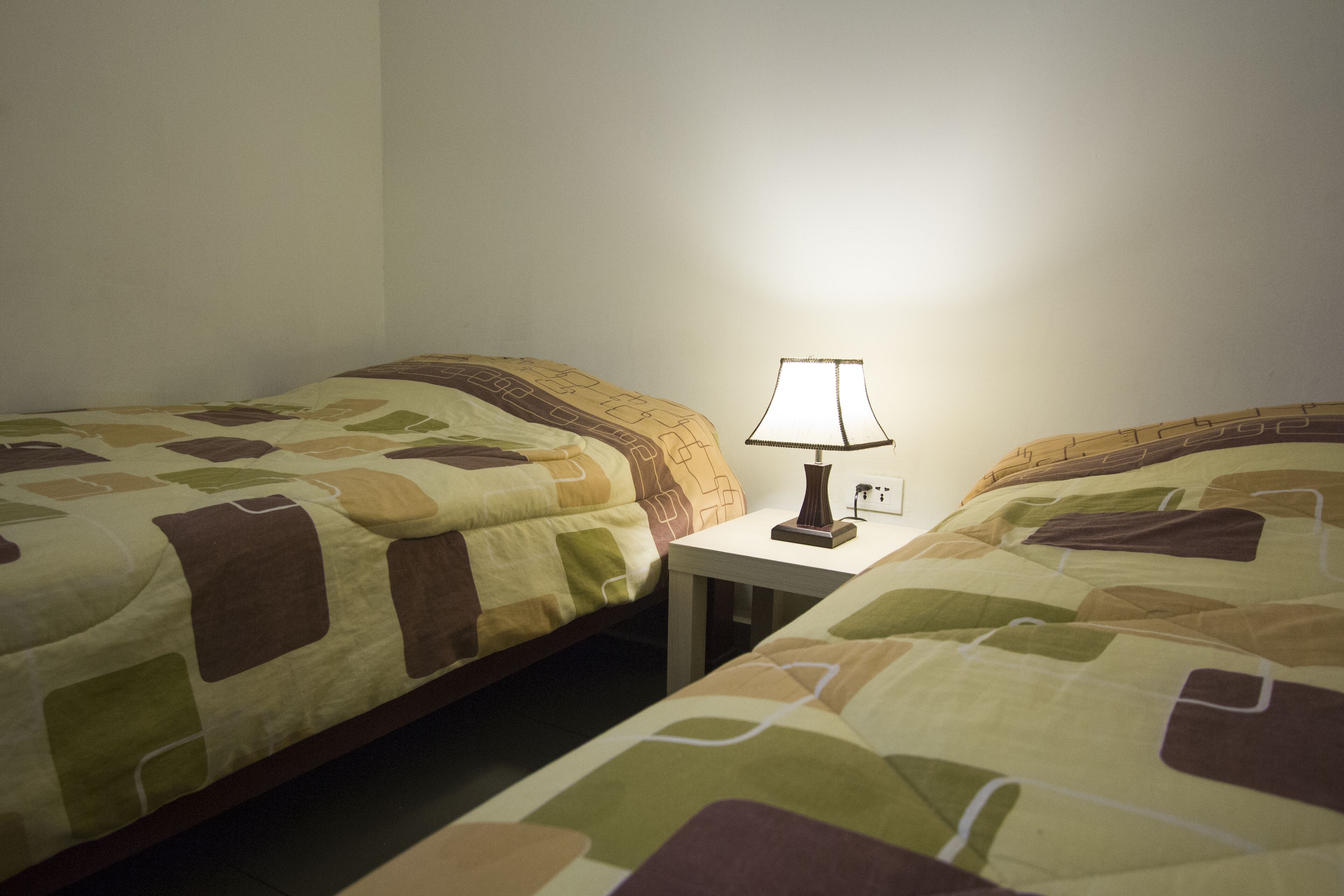 A room with two beds, each with a patterned bedspread, a small bedside table with a lamp, and a wall socket.