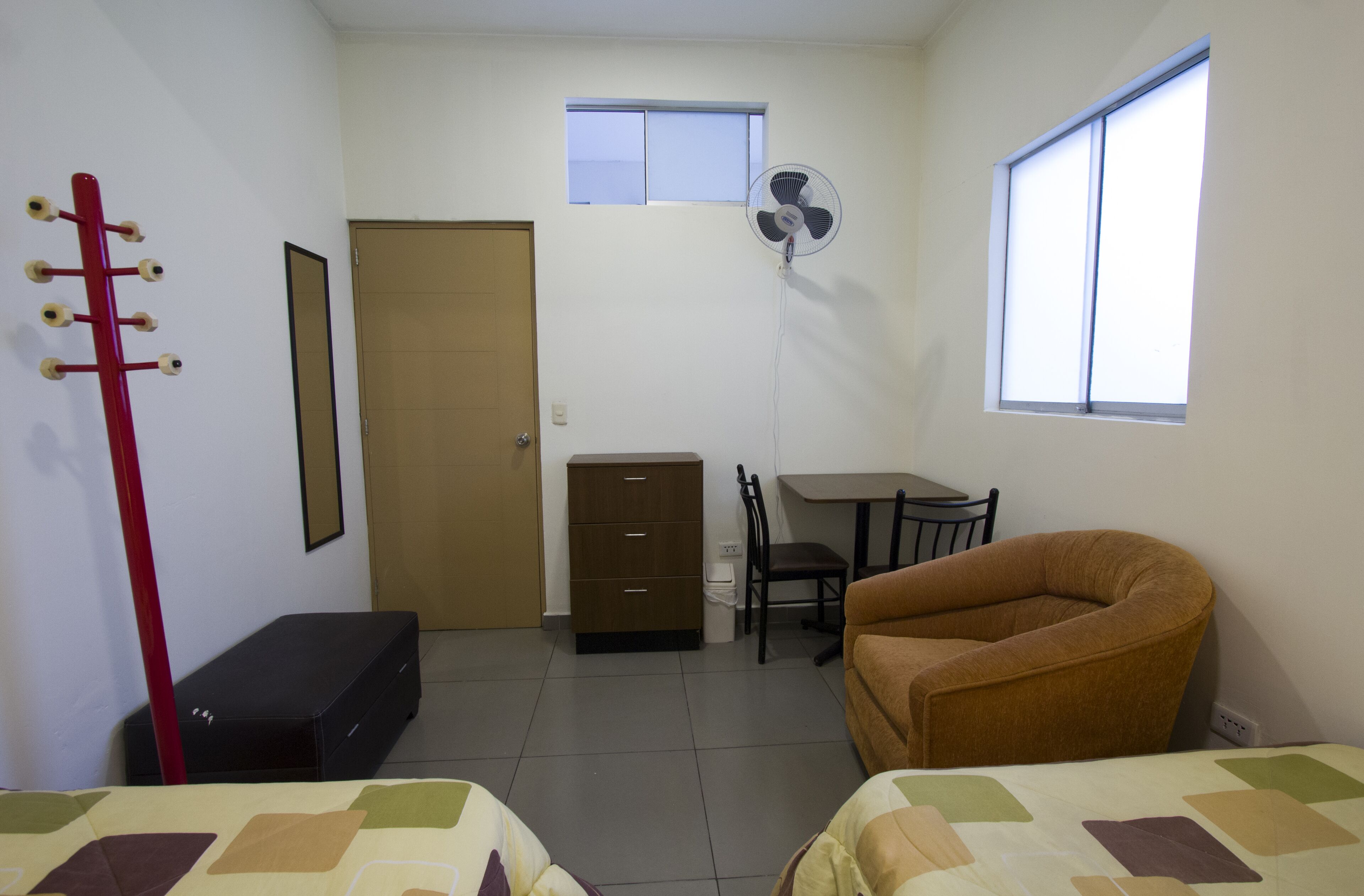 Une petite pièce propre, avec un lit, une chaise, un bureau, une commode, un ventilateur et une fenêtre.