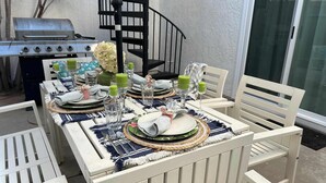 Een eettafel buiten, met witte stoelen, een barbecue en een wenteltrap op de achtergrond.