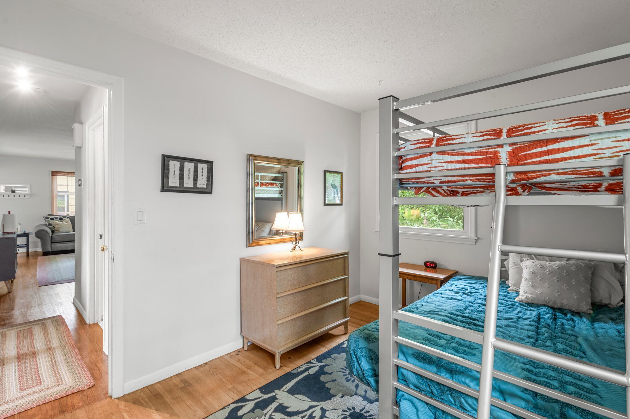 A bedroom with a bunk bed, a dresser, a mirror, and a window with a view of greenery.