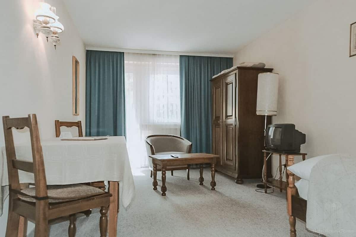Habitación de hotel con una mesa de comedor, sillas, televisor sobre un mueble bajo, armario y ventana con cortinas.
