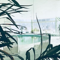An indoor swimming pool with clear glass walls, surrounded by potted plants and white chairs.