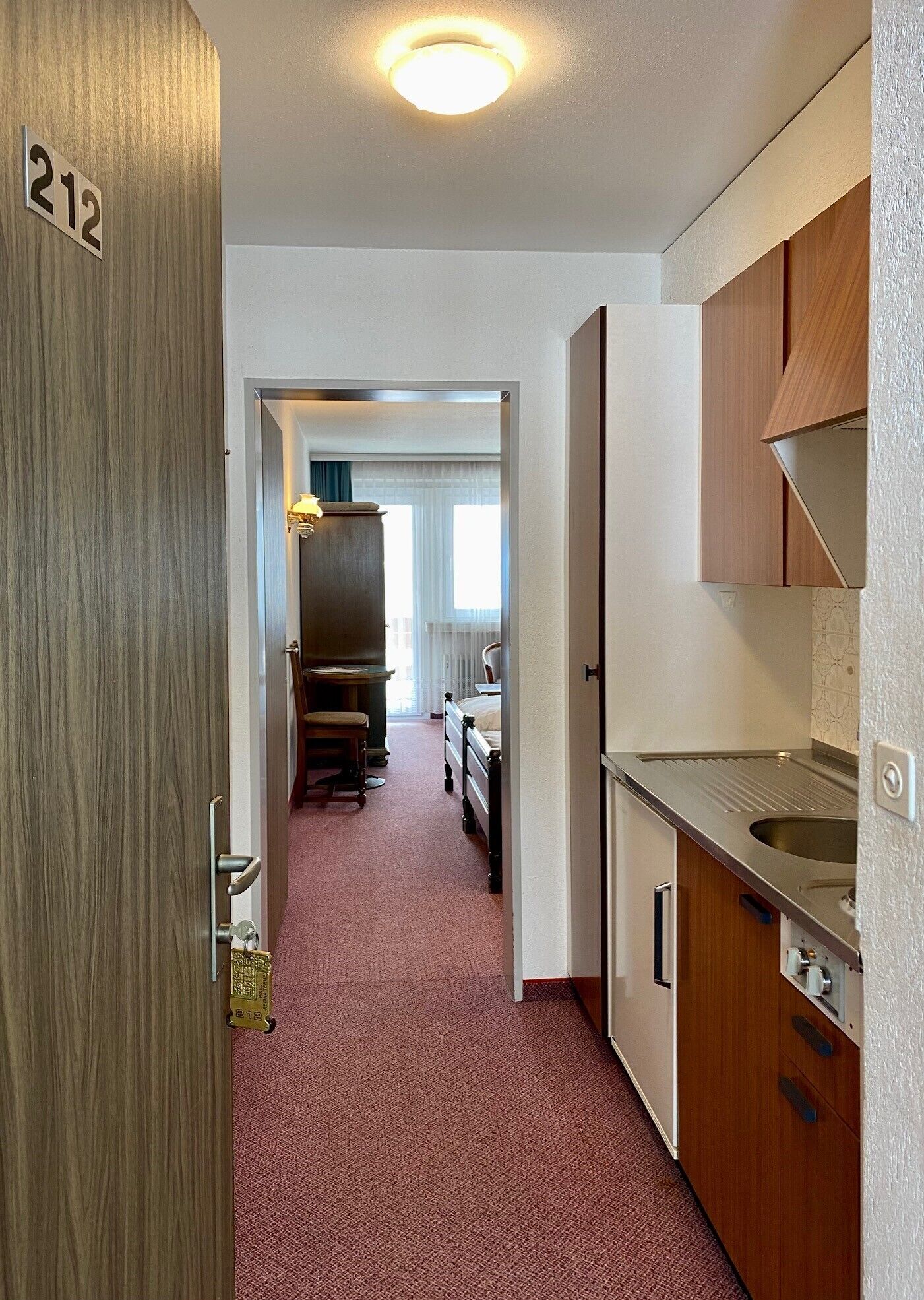 A hotel room with a wooden door marked '212', a kitchenette with a sink and cabinets, a bed with a nightstand, and a hallway leading to a dining area.
