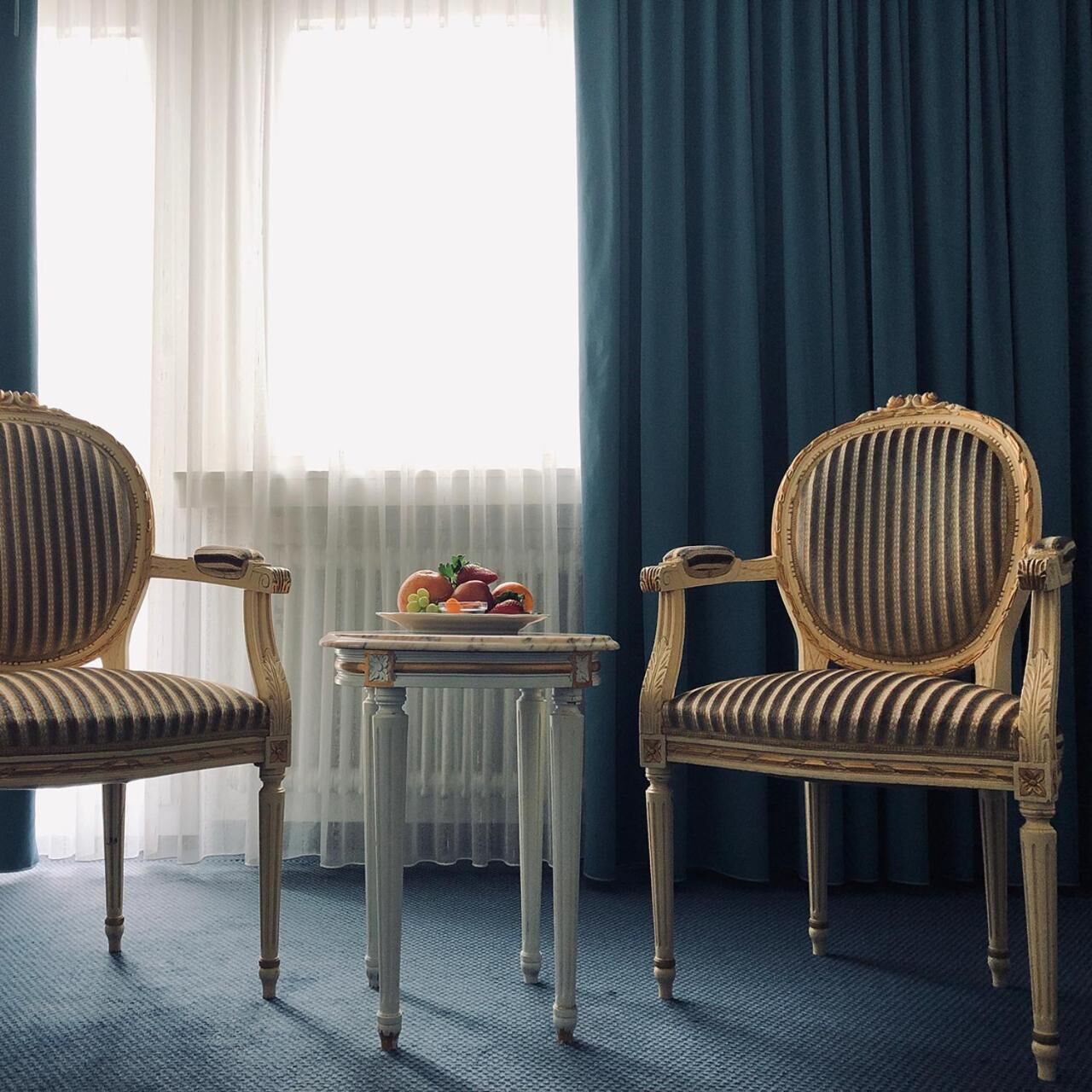 A room with two vintage-style chairs, a small table with a fruit bowl, and a view through sheer curtains.