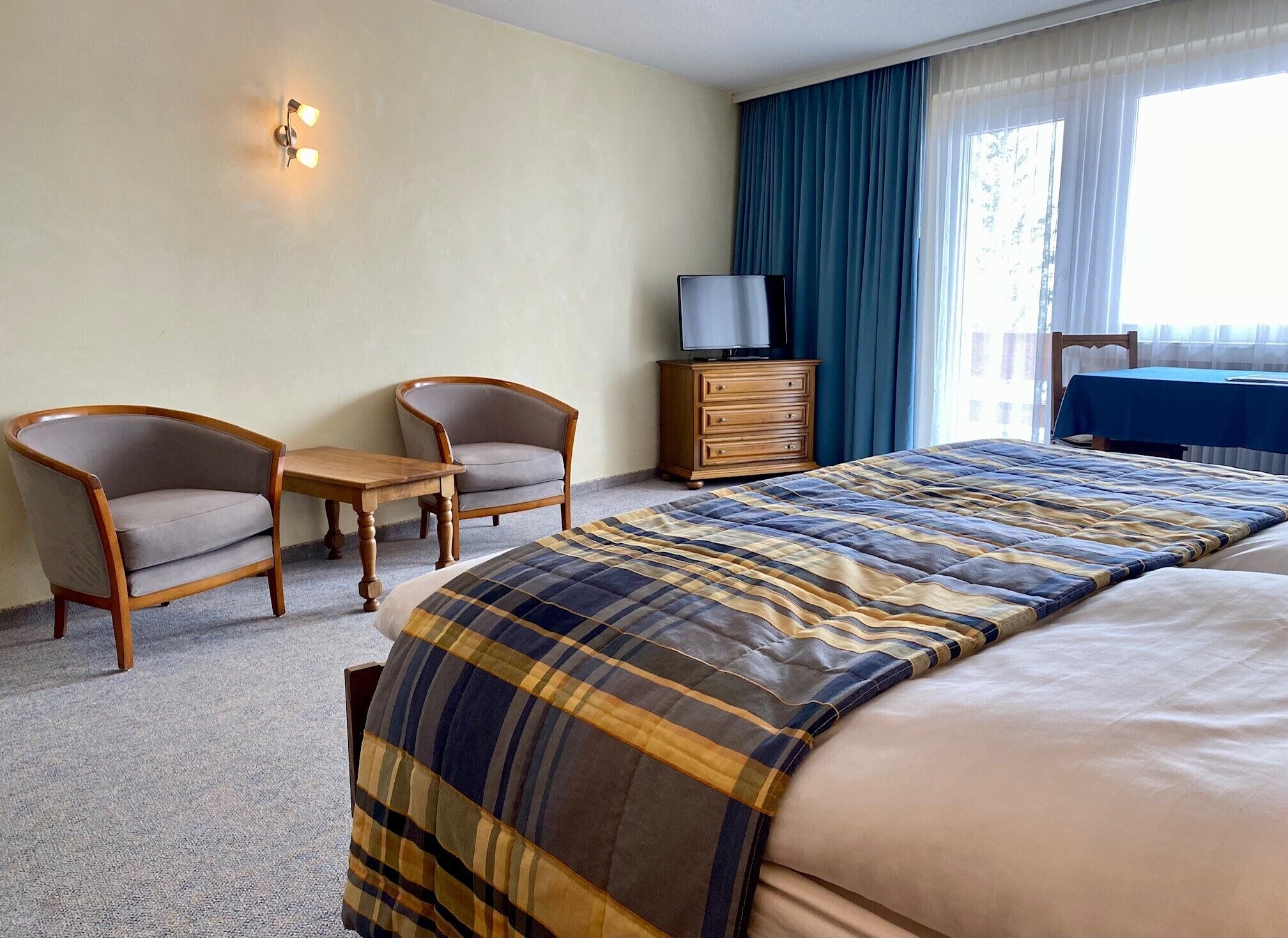 A hotel room with a bed, two armchairs, a small table, a dresser, a television, and a window with curtains.