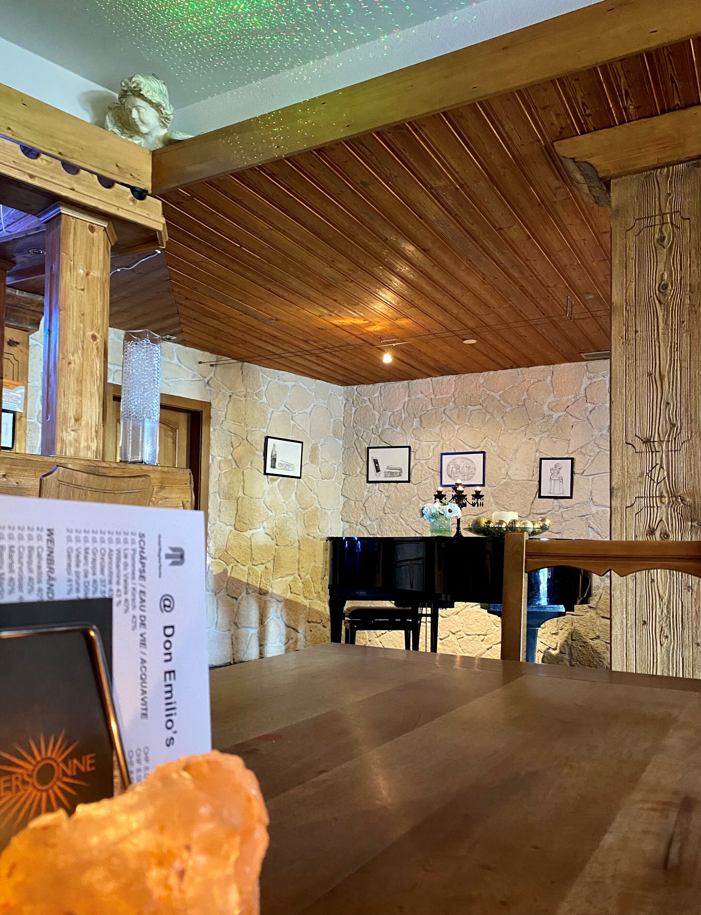 A room with a stone wall, wooden ceiling, and a piano. There are framed pictures on the wall and a table with a laptop and a book.