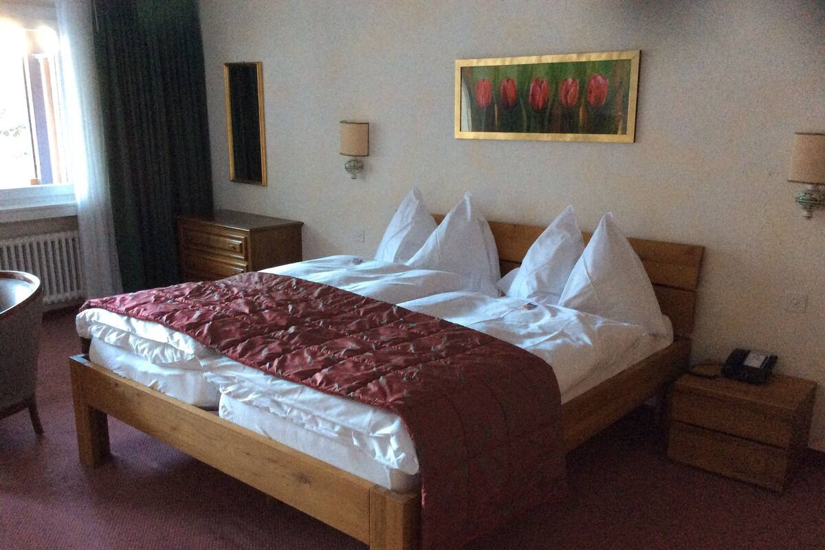 Una cama bien hecha con una colcha rojo y blanco, un cabecero de madera y almohadas blancas. Hay una mesita de noche con una lámpara, un aparador y un cuadro enmarcado con tulipanes rojos en la pared.