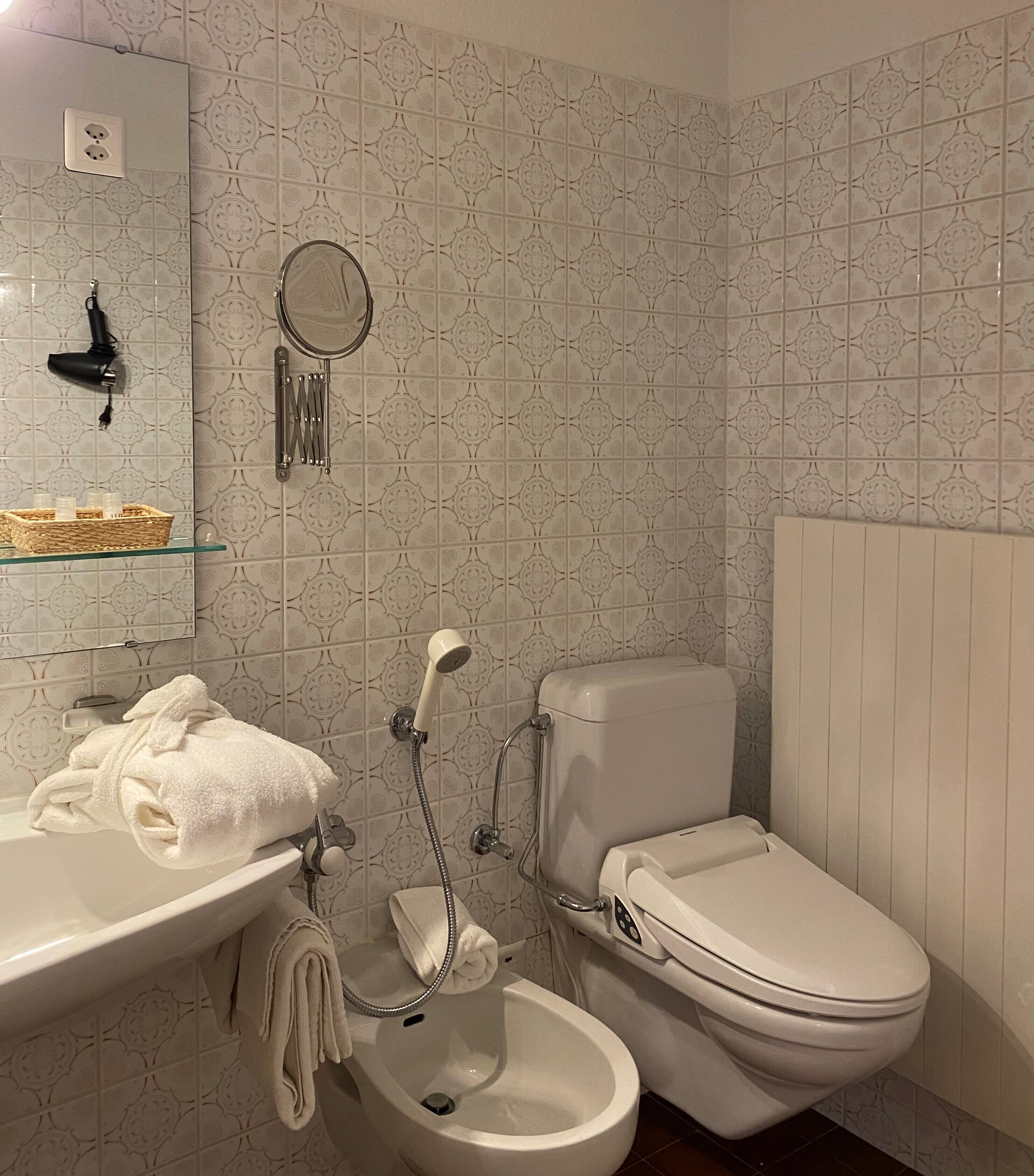 A bathroom with a toilet, bidet, and sink. There is a towel rack with towels, a hairdryer, and a basket on the sink.