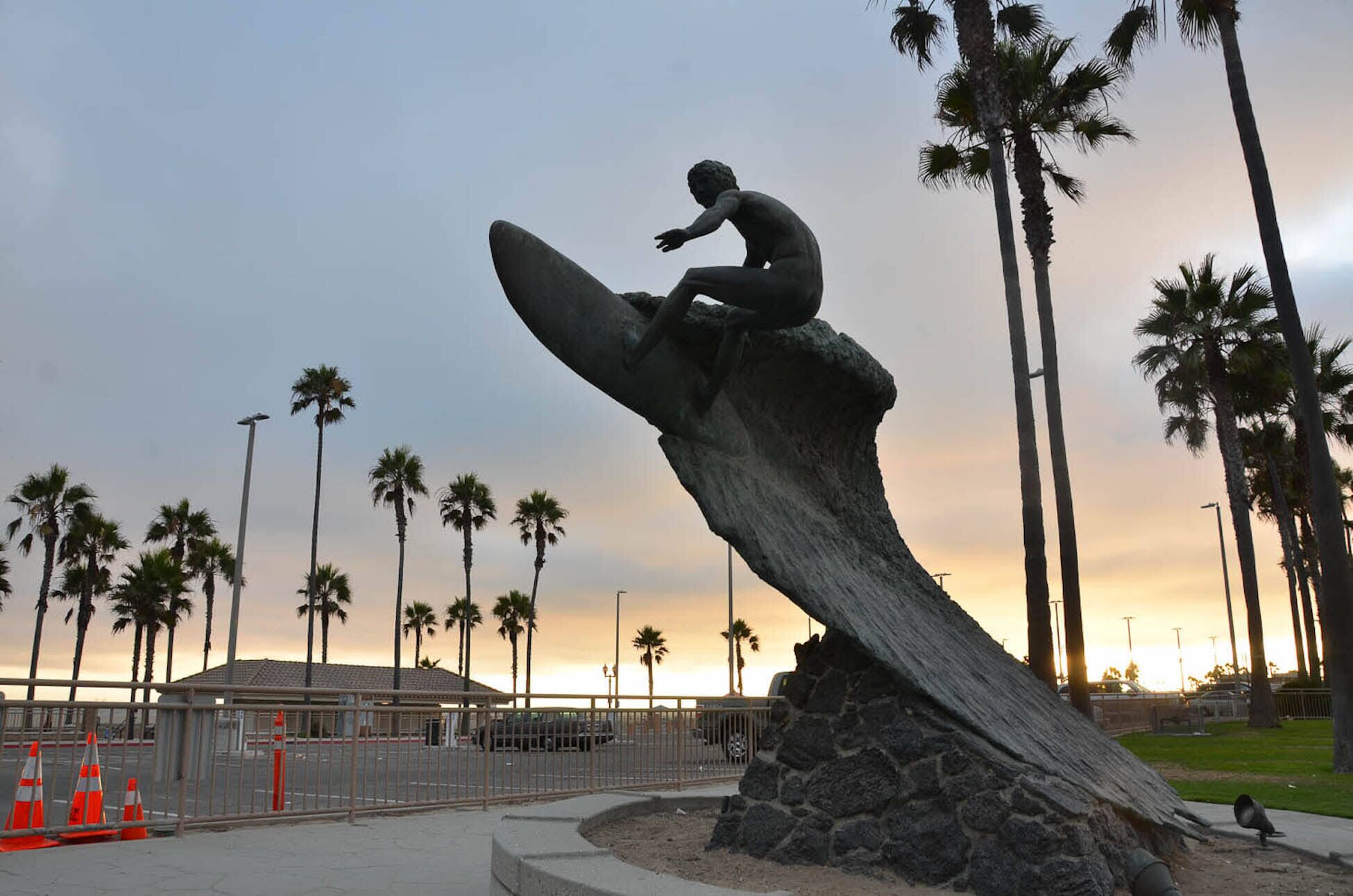 Une sculpture représentant une silhouette en train de faire de la planche à voile sur une vague, entourée de palmiers et avec un coucher de soleil en arrière-plan.