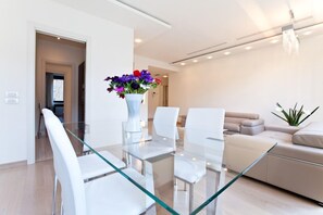 A modern living room with a glass dining table, white chairs, a beige sofa, and a vase of flowers.