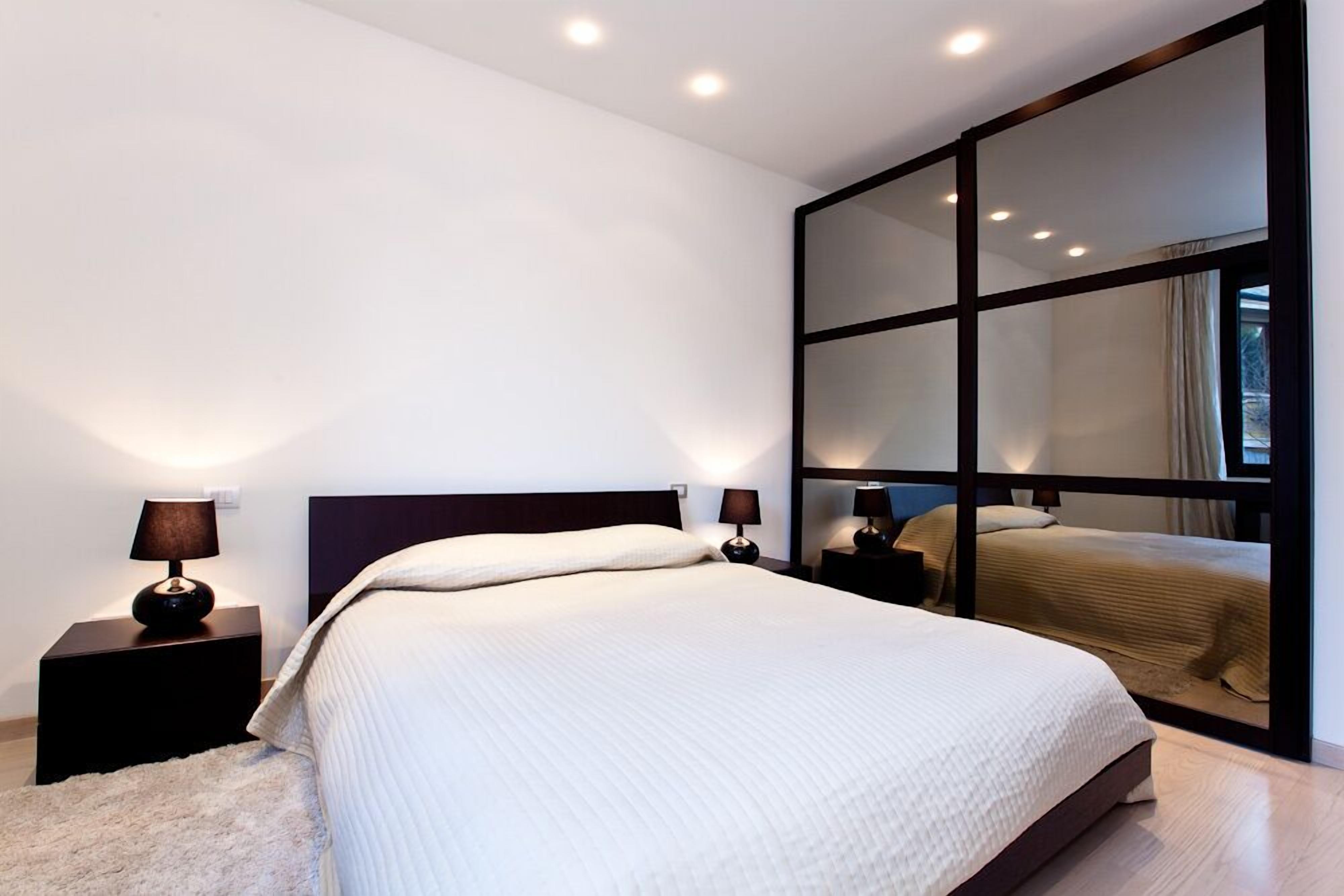 Een moderne slaapkamer met een groot bed, twee nachtkastjes met lampen en een grote spiegel.