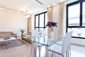 Un salon moderne avec une table à manger en verre, des chaises blanches, un canapé beige et une grande fenêtre avec des rideaux.