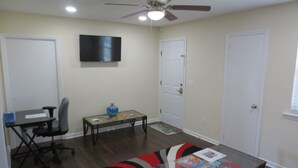 A living room with a ceiling fan, a flat-screen TV, a glass coffee table with magazines, a black office chair, and a red and black rug. - Apartment In Downtown Durham (Durham)