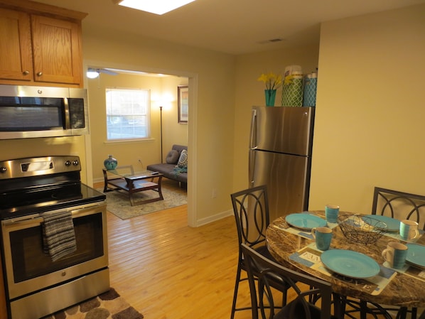 A compact kitchen with a microwave, oven, and refrigerator. There is a dining table with blue plates and a vase with yellow flowers. A sofa and a small rug are in the living area.