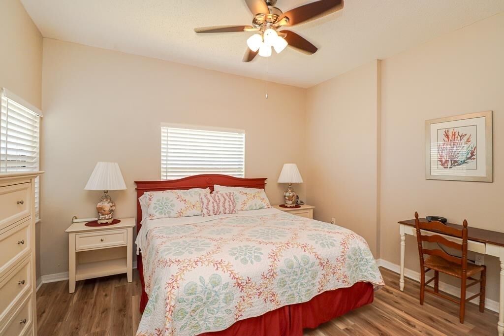 A bedroom with a bed, two bedside tables with lamps, a desk with a chair, a ceiling fan, and a window with blinds.