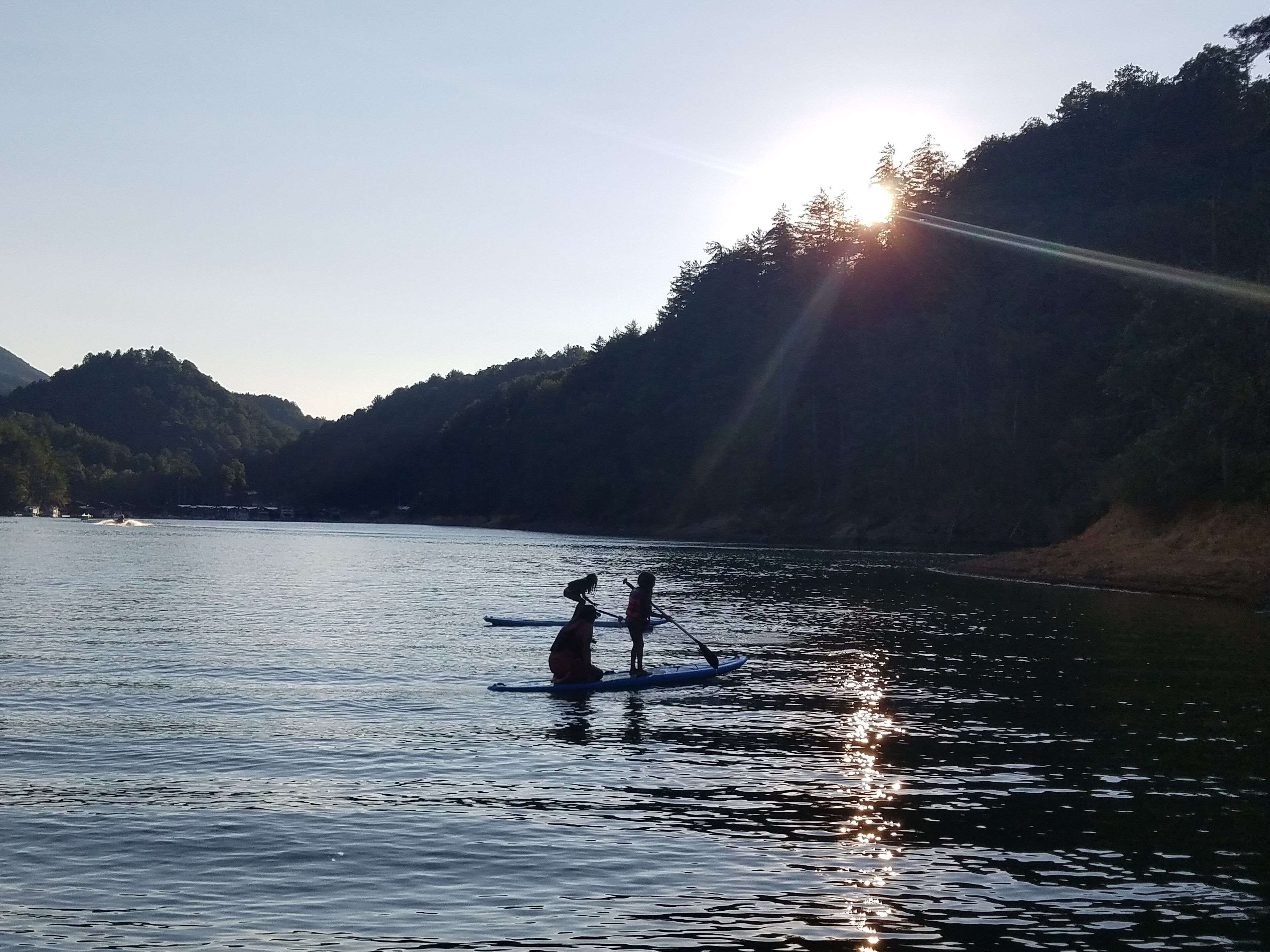 Two people paddleboarding on a calm lake with a forested hillside in the background and the sun setting behind them.