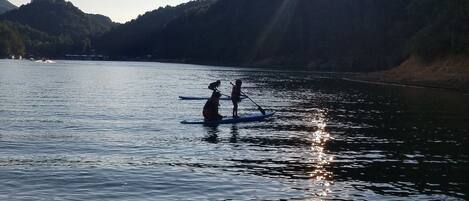Two people paddleboarding on a calm lake with a forested hillside in the background and the sun setting behind them.
