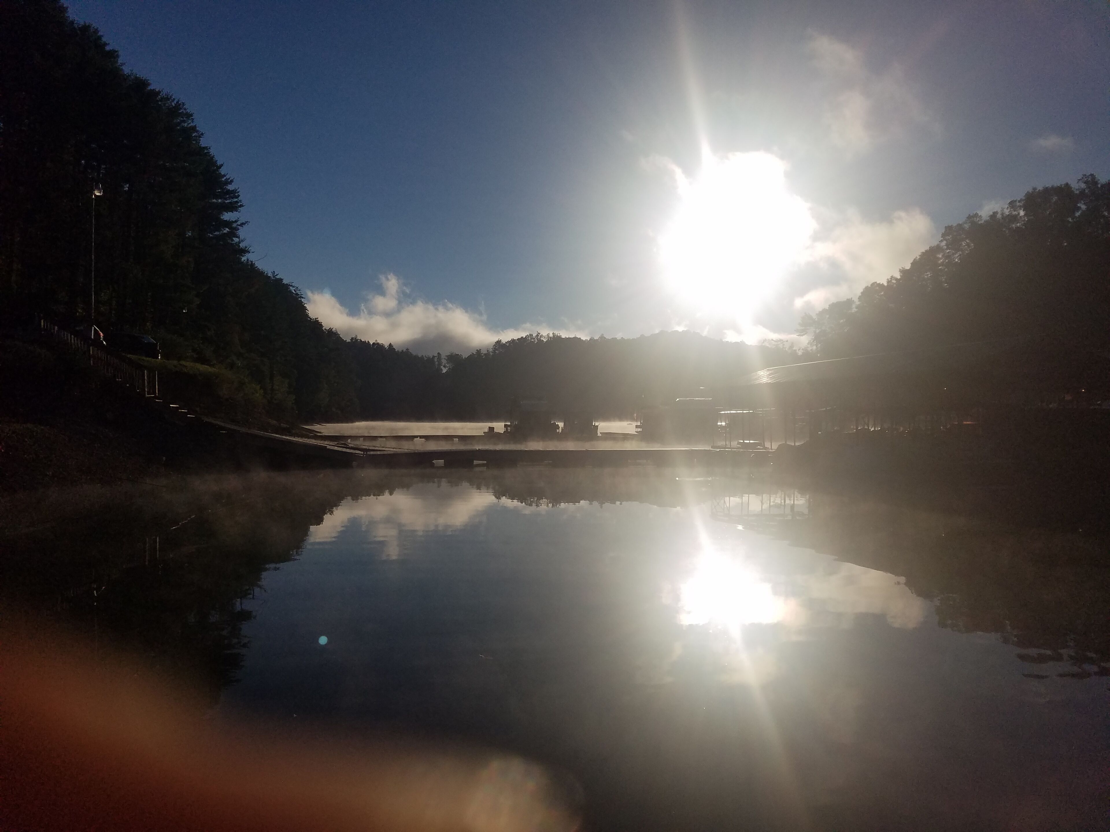 A serene lake with a clear reflection of the sun and surrounding trees.