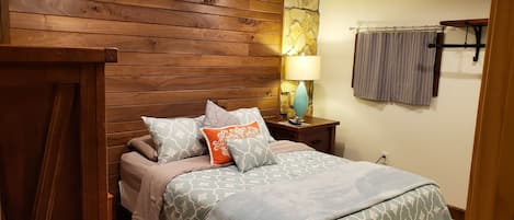 A bedroom with a wooden headboard, a bed with patterned bedding, a nightstand with a lamp, and a curtain on the window.