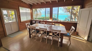 Un coin repas en bois avec une grande table et des chaises, de grandes fenêtres donnant sur la mer et un ventilateur de plafond.