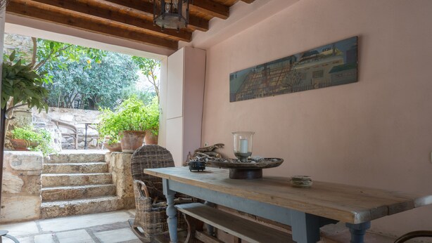 A rustic dining area with a wooden table, bench, and wicker chairs. A stone staircase leads to an outdoor area with potted plants and a view of trees. - Acropolis Villa (Athens)